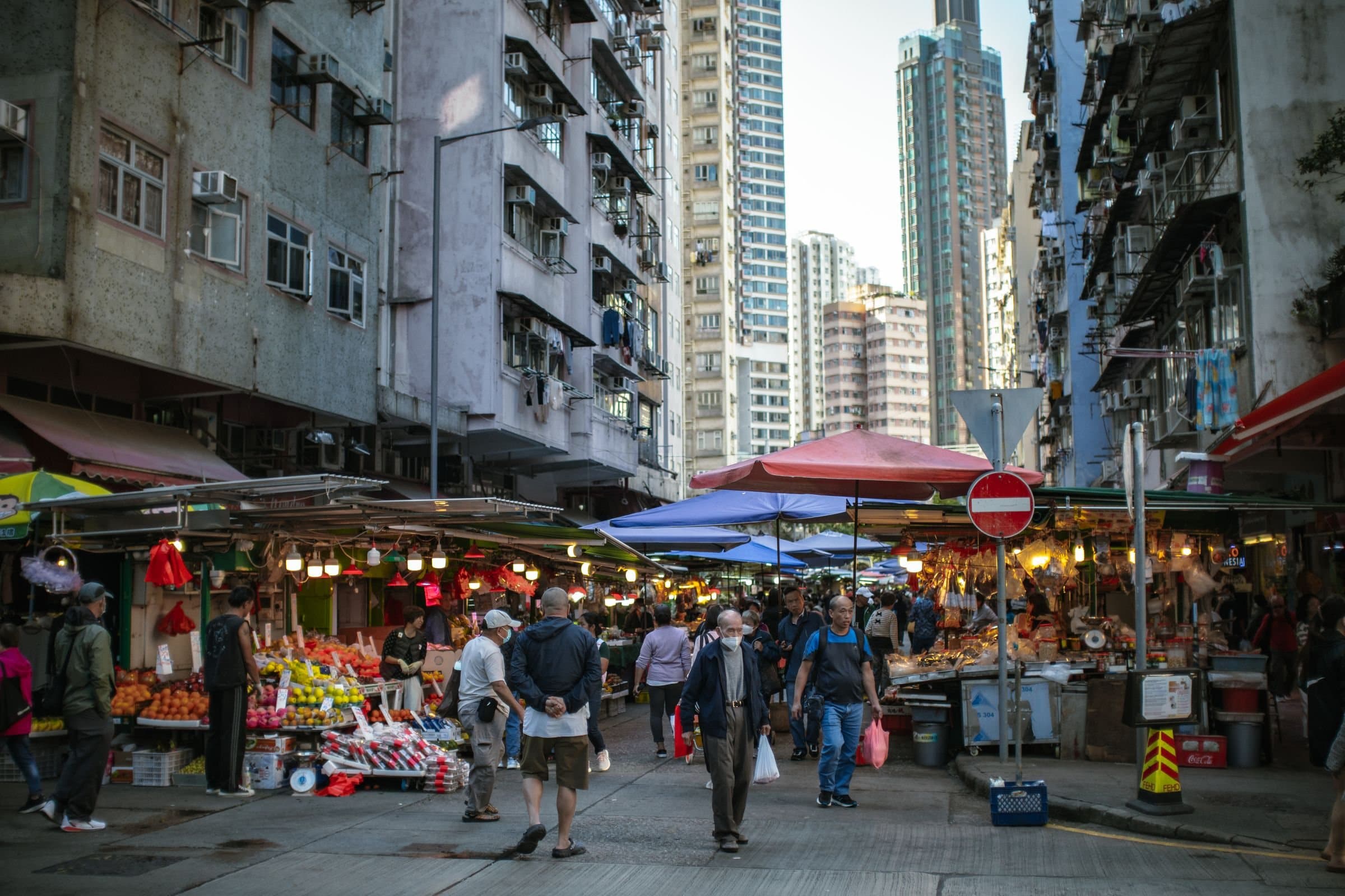 Hong Kong street market with fruit stalls under red and blue umbrellas, shoppers walking between old tenements and towers