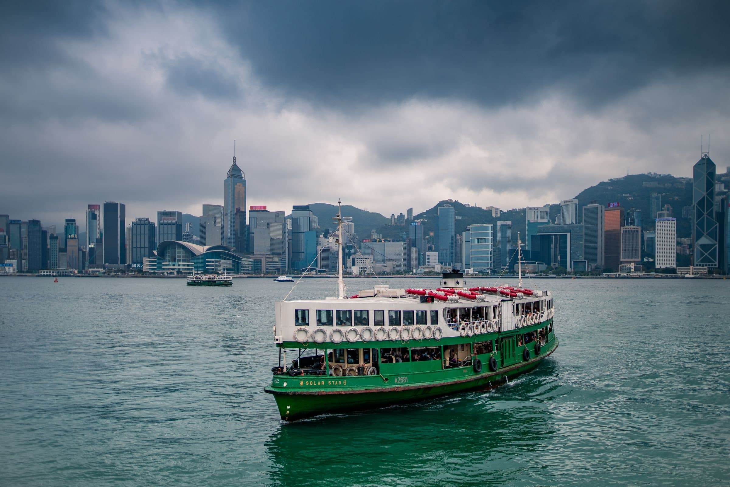 Green and white Star Ferry crossing Victoria Harbour, with the Hong Kong Island skyline under heavy grey clouds